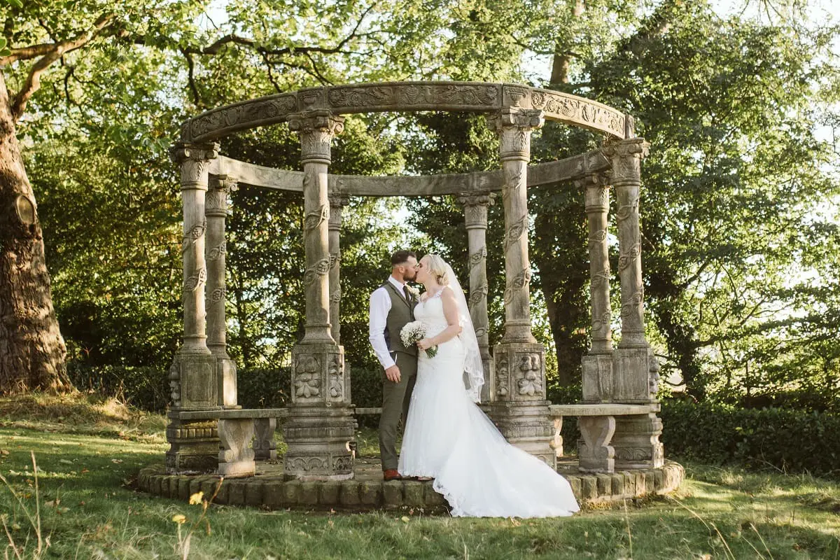 A newlywed couple stands embracing inside a decorative stone gazebo in a lush garden setting. The bride is in a white gown, and the groom is in a suit.