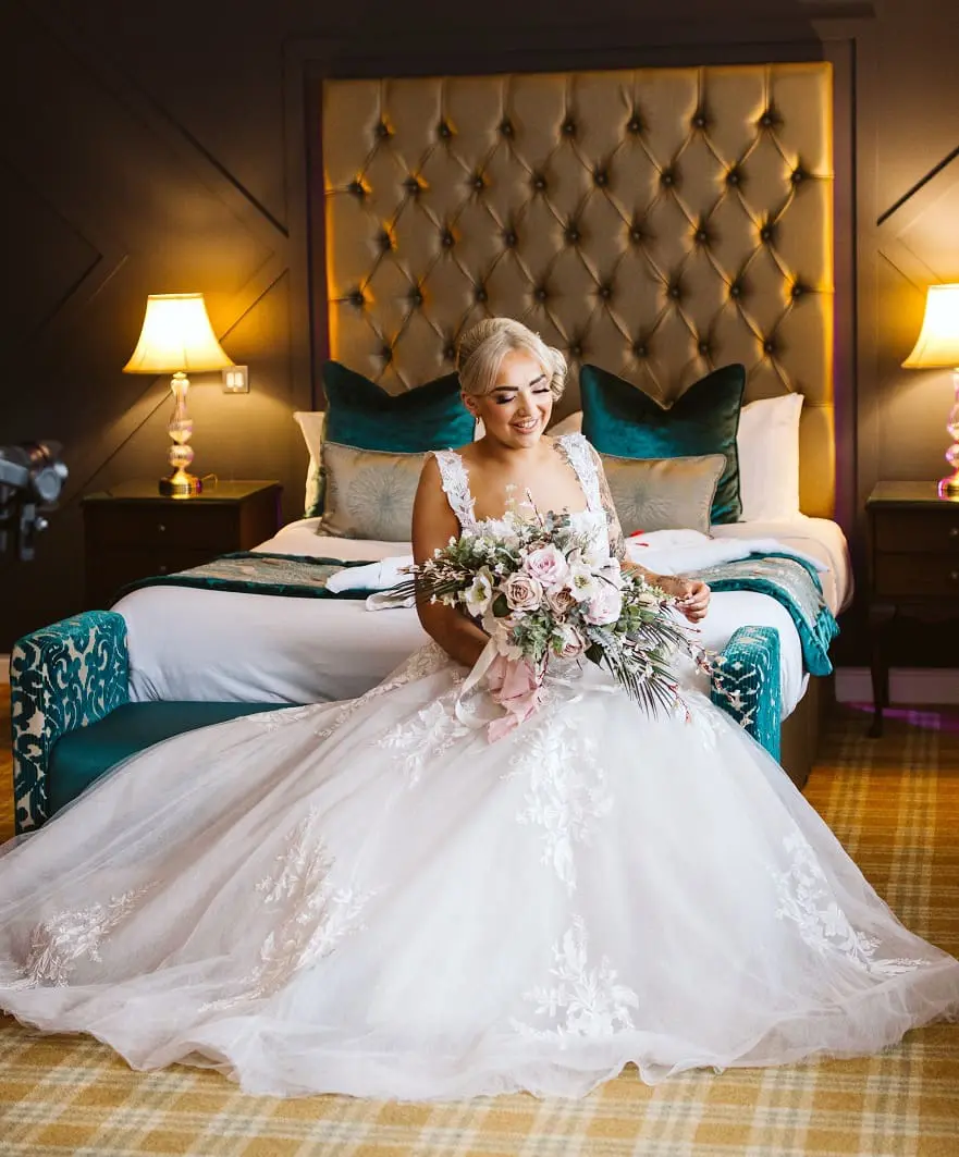 A bride in a white gown sits on the edge of a teal and gold-themed bed, holding a bouquet, with two lit lamps on the nightstands behind her.