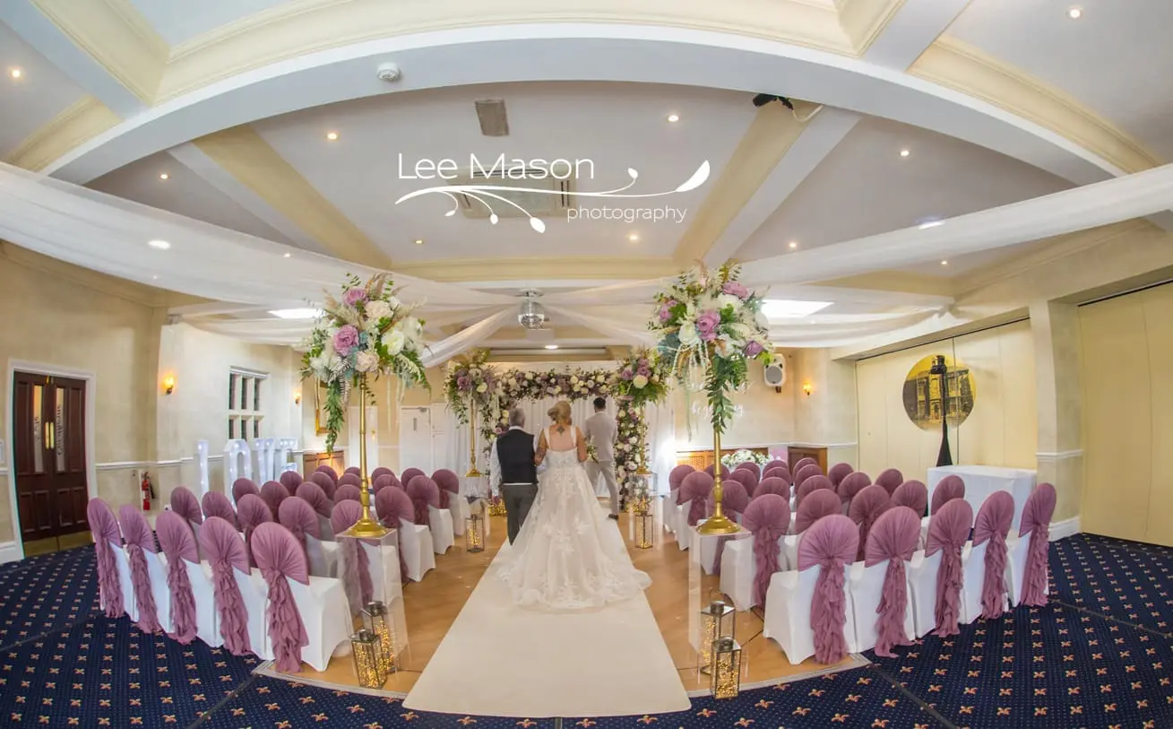A bride and groom stand at the altar in a decorated wedding venue with rows of purple chair covers and floral arrangements. "Lee Mason Photography" is visible on the image.