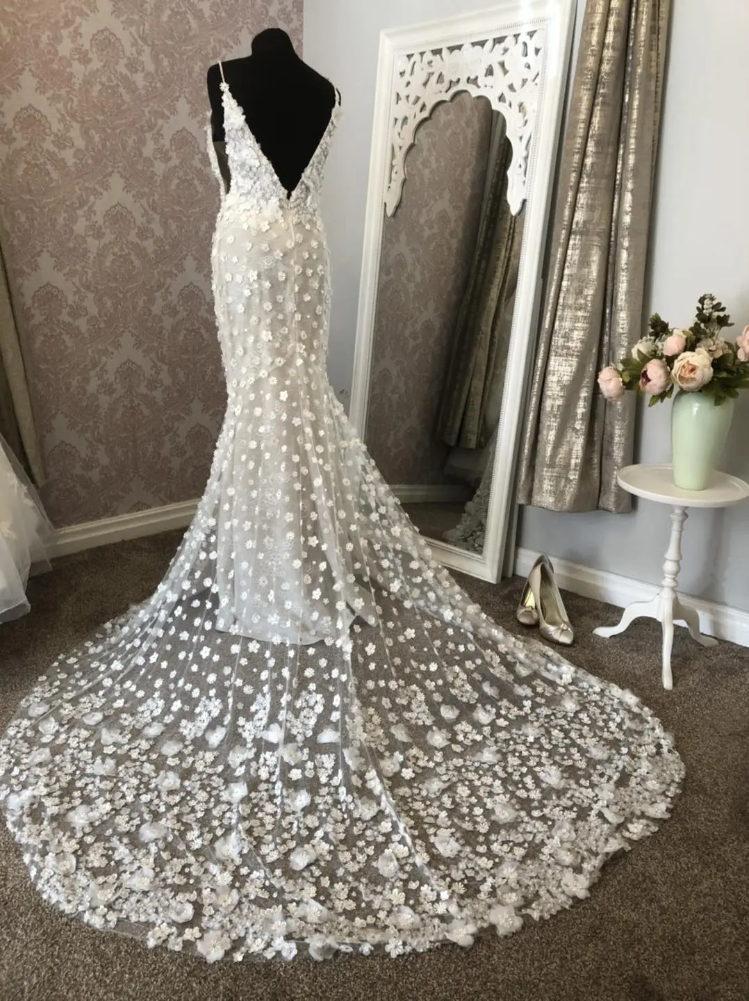 A white lace wedding dress with floral patterns graces a mannequin, its long train elegantly flowing. Nearby, a mirror reflects gold shoes and a small table adorned with roses, capturing the essence of an exquisite wedding planning scene in the UK.