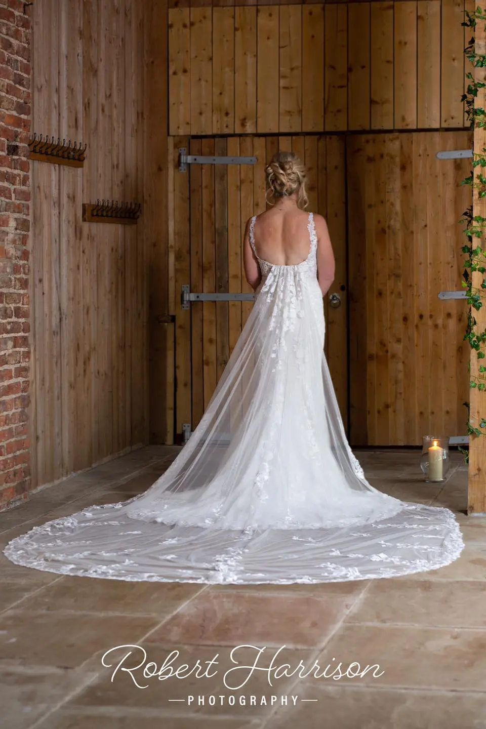 A bride in a white backless wedding gown stands facing a wooden wall with a lit candle nearby. The dress has a long train spread out on the floor. Text at the bottom reads "Robert Harrison Photography.