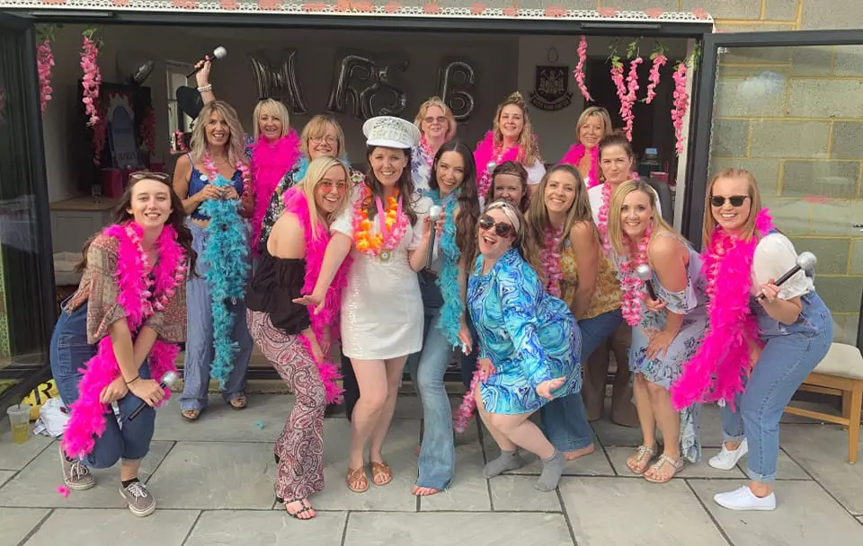 A group of women, some wearing feather boas, are gathered together outside in front of a building for a celebration. One woman in the center wears a white outfit and hat, and all are smiling at the camera.