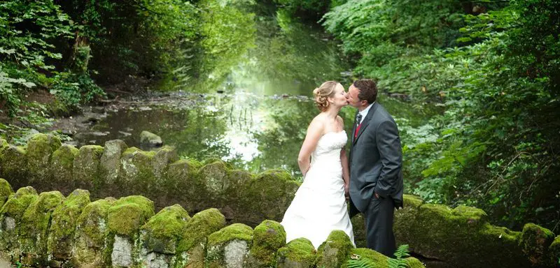 A bride and groom sharing a tender kiss on a stone bridge in a picturesque wooded area for their romantic wedding ceremony.