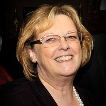 A woman wearing glasses and pearls smiles for the camera while working on her wedding planning business.
