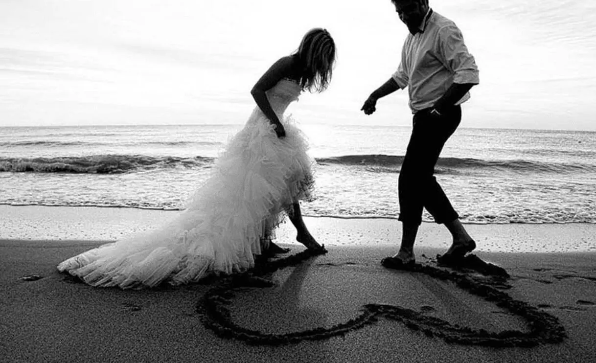 A bride and groom standing on the beach with a heart drawn in the sand at their wedding in UK.