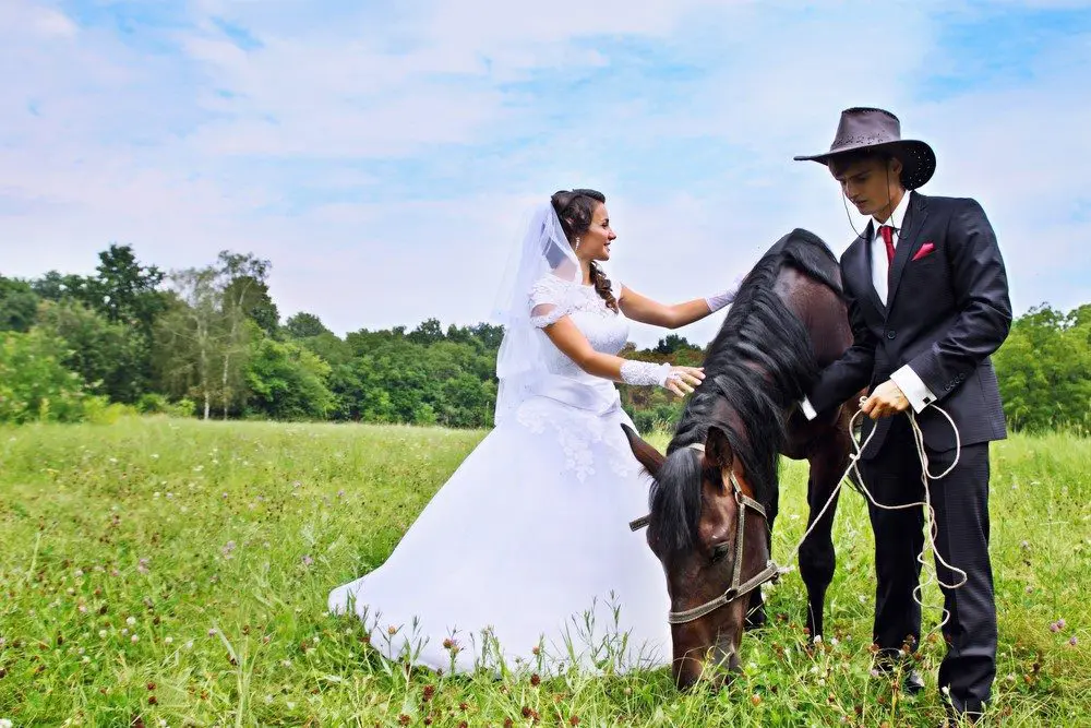 A bride and groom standing next to a horse in a field, capturing the essence of their wedding day.