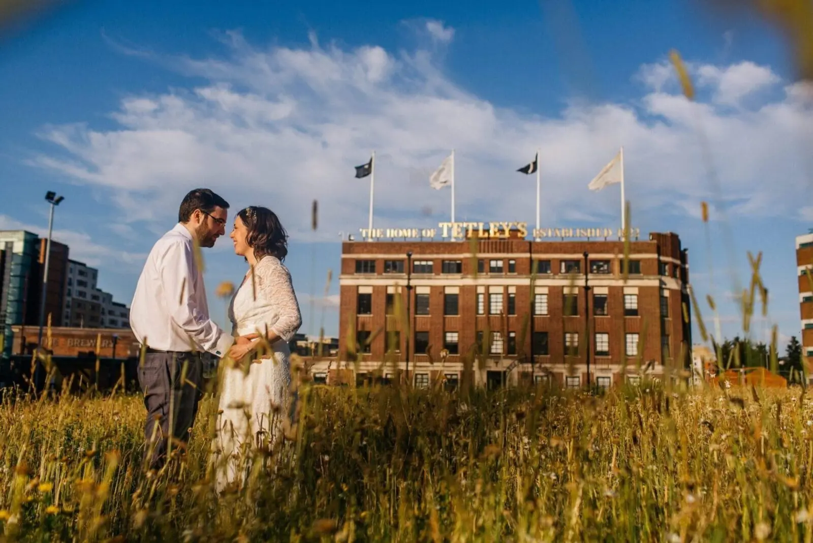 A wedding couple standing in tall grass in front of a building, beautifully captured for their wedding album.