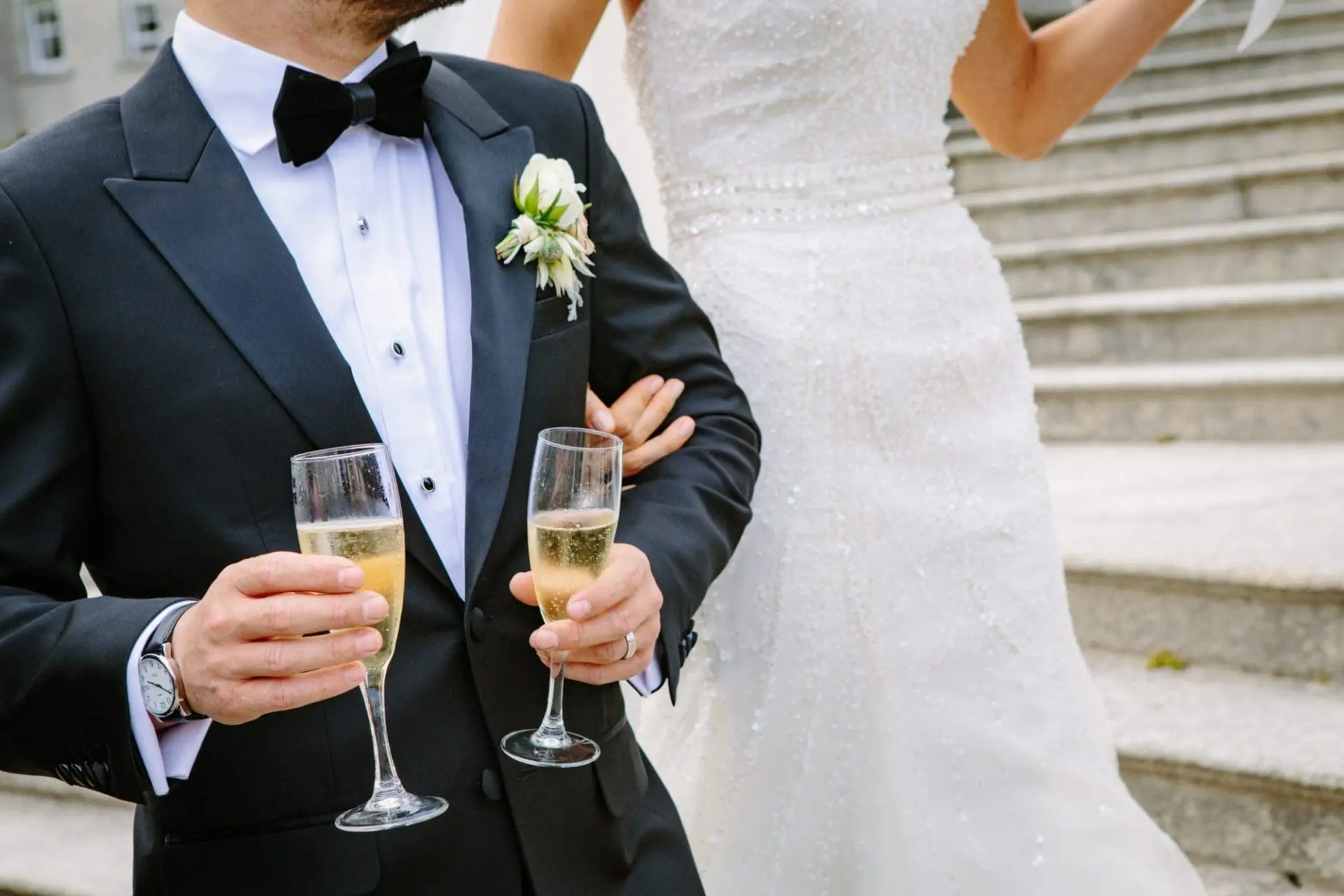 A bride and groom, elegantly dressed, hold champagne glasses on the steps of a grand venue as they celebrate their marriage.