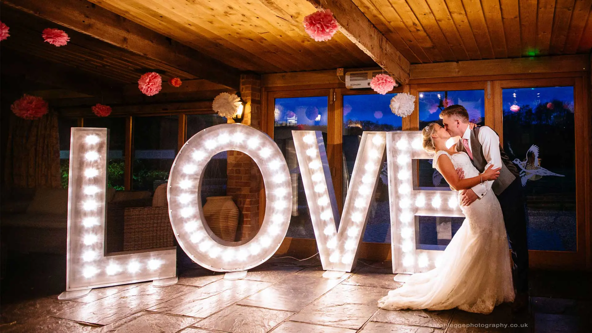 A bride and groom share a romantic kiss in front of lighted love letters as part of their wedding planning journey.