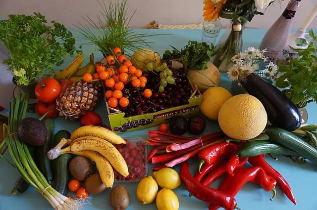 A table filled with various fruits and vegetables, including bananas, peppers, lemons, eggplants, berries, and melons, along with a box of cherry tomatoes and leafy greens. Flowers and a bottle are in the background, reminiscent of a beautifully curated wedding planning spread.