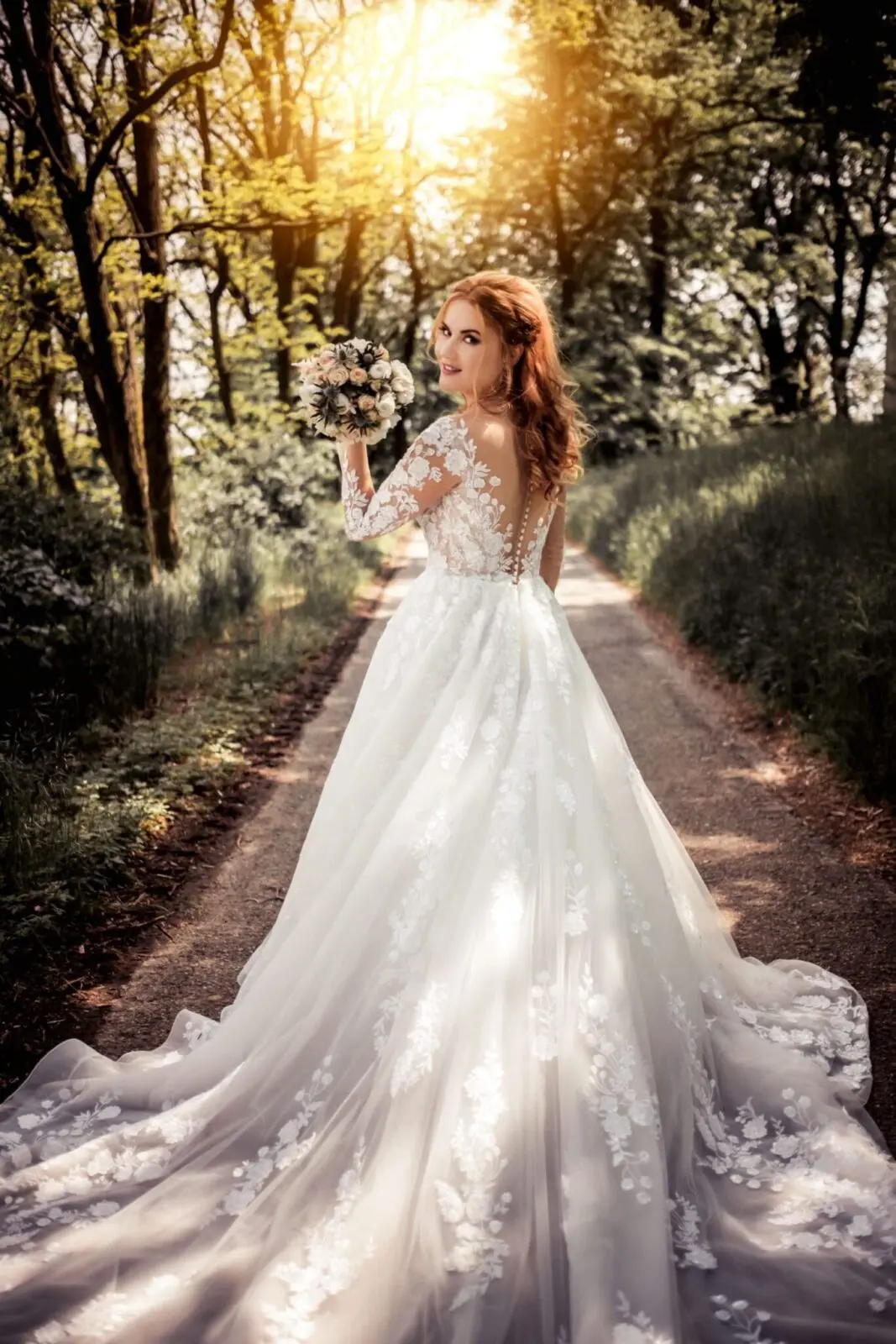 A bride with red hair in a long white lace wedding dress holds a bouquet and looks back while walking down a tree-lined path at sunset, the epitome of timeless elegance and careful wedding planning.
