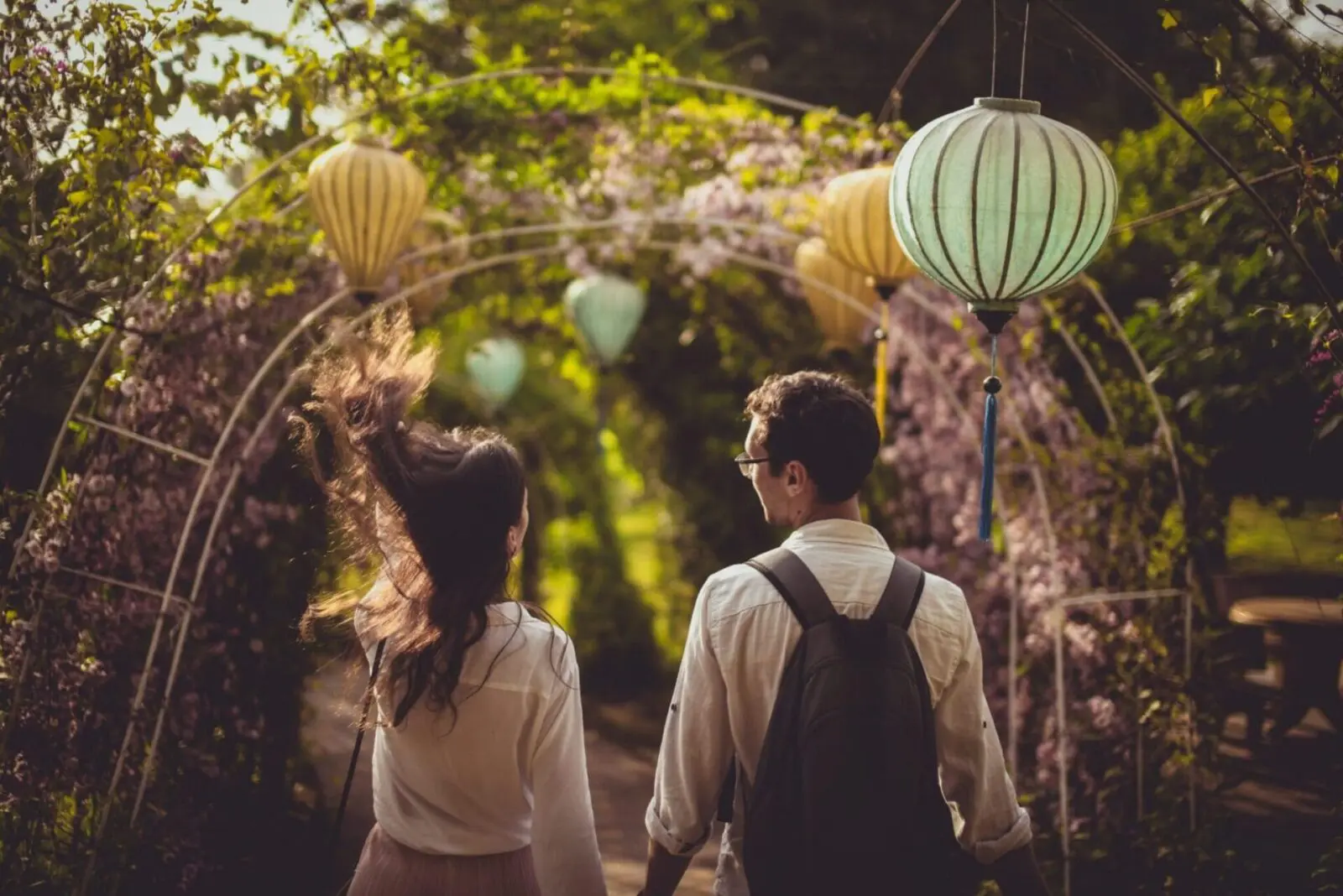 A couple walks under an archway decorated with hanging lanterns and surrounded by lush greenery, as if stepping straight out of a meticulously planned wedding dream.