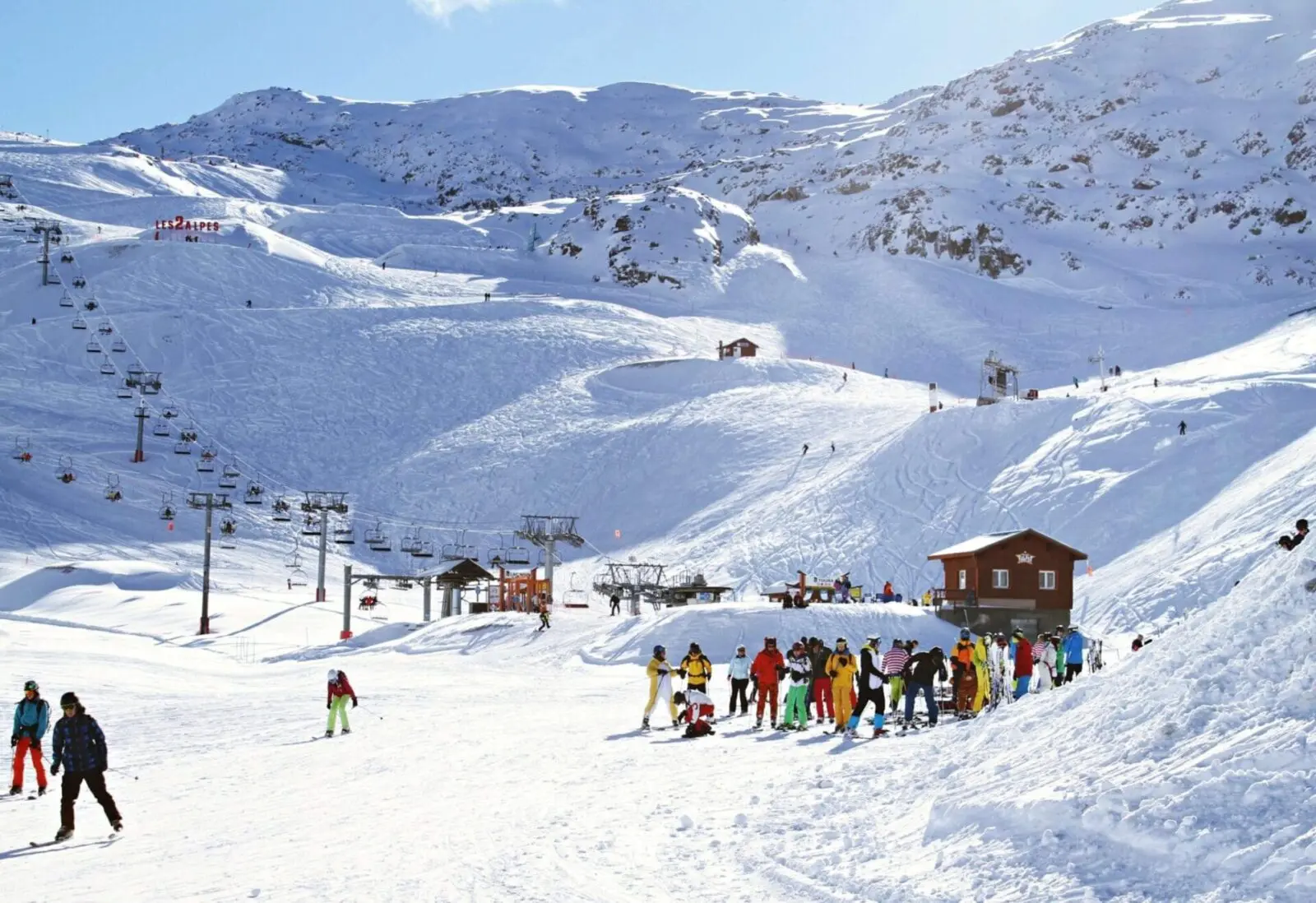Skiers gather at the base of a snowy ski slope with chair lifts and a small building visible. Snow-covered mountains and a clear blue sky form the backdrop, making it an ideal location for wedding services amidst nature's beauty.