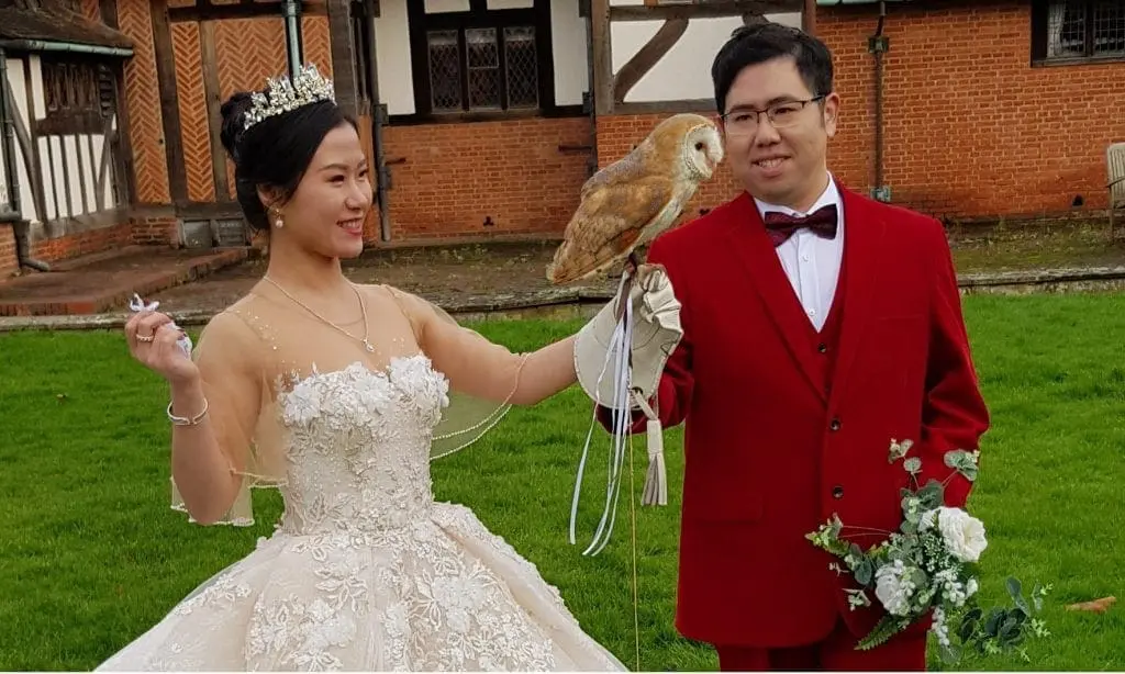 A bride in a white gown and crown holds a barn owl on her gloved hand, standing next to a groom in a red suit holding flowers. They are outdoors near a half-timbered building, celebrating their wedding in the picturesque UK countryside.