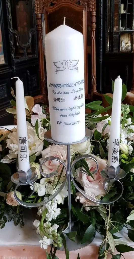 A decorative display with a large white candle and two smaller white candles on either side, surrounded by flowers. The larger candle features congratulatory text, two doves, and the date "2nd June 2019," perfect for wedding services in the UK.