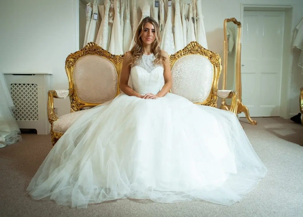 A woman in a white wedding dress sits on an ornate, gold-trimmed sofa in a bridal shop, surrounded by other wedding dresses hanging on racks, contemplating the next steps of her wedding planning.