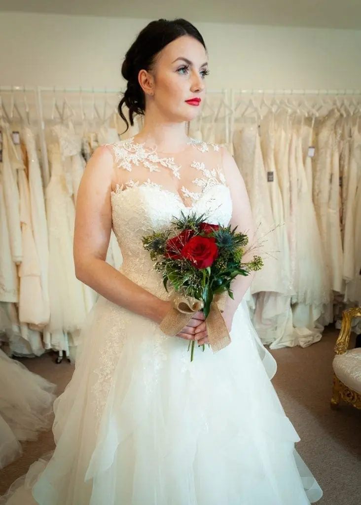 A bride in a white lace wedding dress holds a bouquet of red roses and greenery, standing in a bridal shop where the wedding business thrives with an array of stunning dresses displayed on hangers in the background.