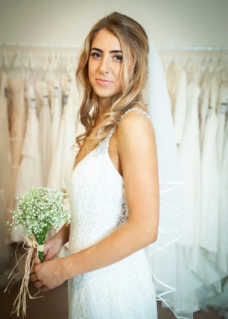 A bride in a white lace wedding dress and veil holds a small bouquet of white baby's breath flowers, standing in front of a rack of hanging wedding dresses—perfect for those deep into wedding planning in the UK.