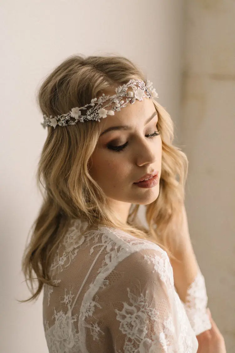 A woman with wavy blonde hair wears a white lace dress and a floral headpiece, looking down thoughtfully, perhaps lost in the details of planning her wedding.