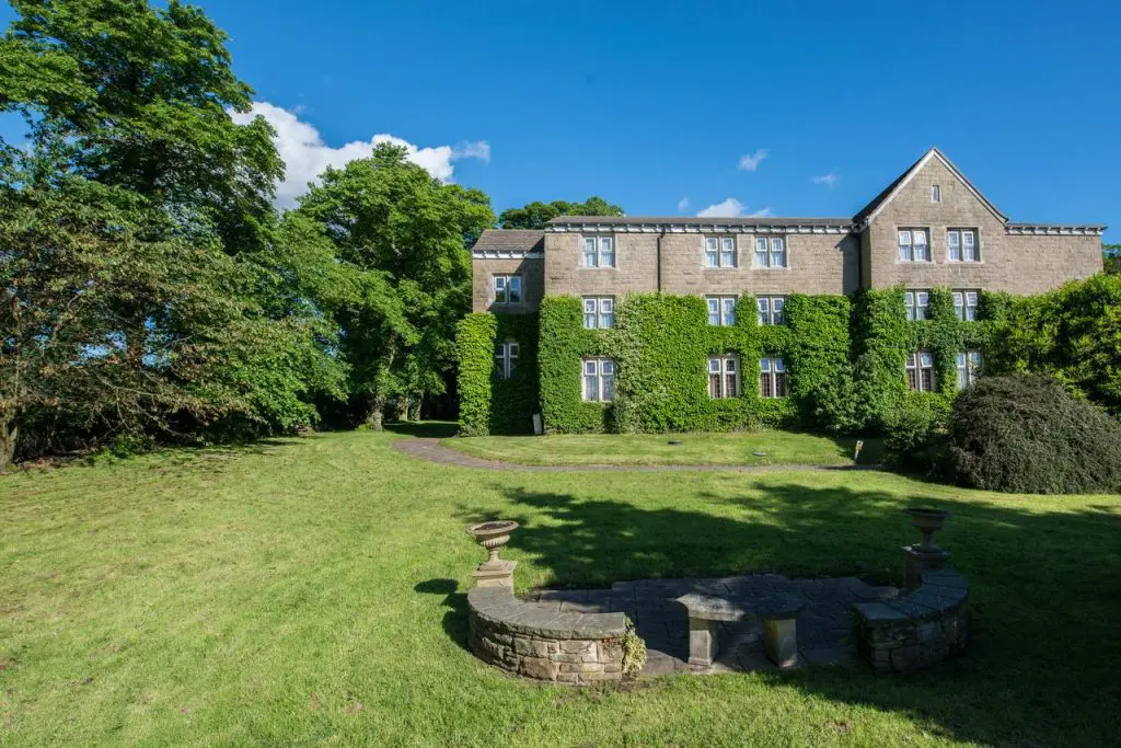 A large stone building covered in ivy, situated in a spacious grassy area with trees, under a clear blue sky. A round stone bench is in the foreground—an idyllic setting perfect for wedding planning in the UK.