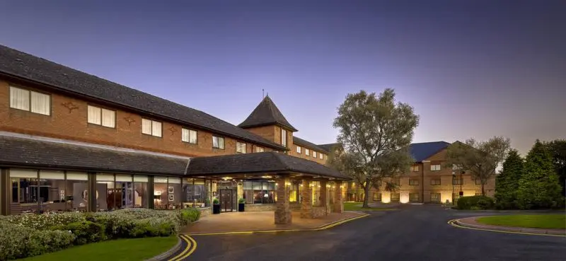 Exterior view of a large red-brick hotel at dusk, featuring a central entrance with a pitched roof and surrounding trees and landscaping, providing an ideal setting for wedding services.