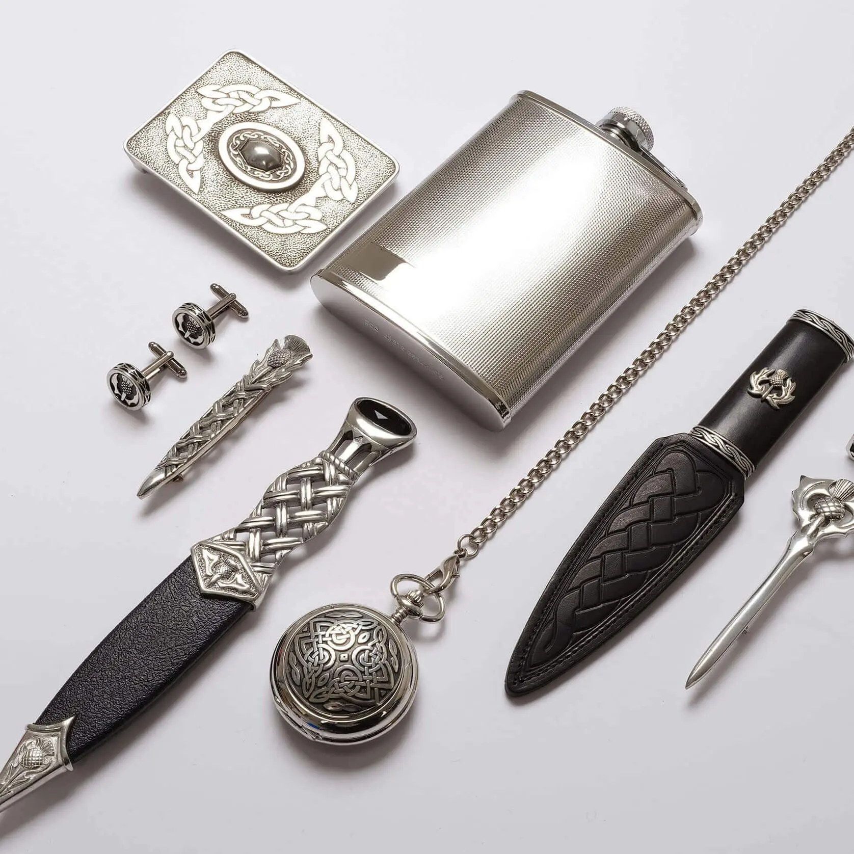 A collection of items including a flask, belt buckle, tie clip, cufflinks, pocket watch, and two small daggers, arranged neatly on a white surface.