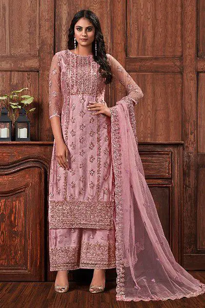 A woman stands in front of a wooden wall, wearing an embellished, pink traditional outfit with a matching sheer dupatta draped over her arm. She has long, dark hair and is wearing earrings.