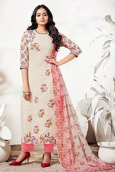 A woman stands posing in a floral-patterned, long-sleeved traditional outfit with matching pants and a flowing draped scarf. She is wearing red heels and is surrounded by white potted plants.