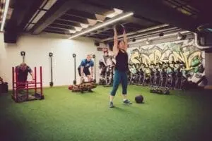 A group of people exercising in a gym with various equipment; one person lifts a medicine ball overhead, another pushes a weighted sled, and two others work with kettlebells in front of graffiti-covered walls.