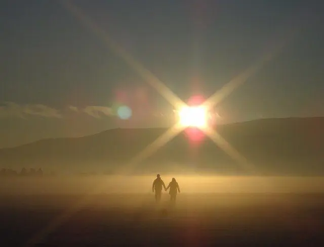 A couple holds hands while walking towards the rising or setting sun in a misty landscape with mountains in the background.