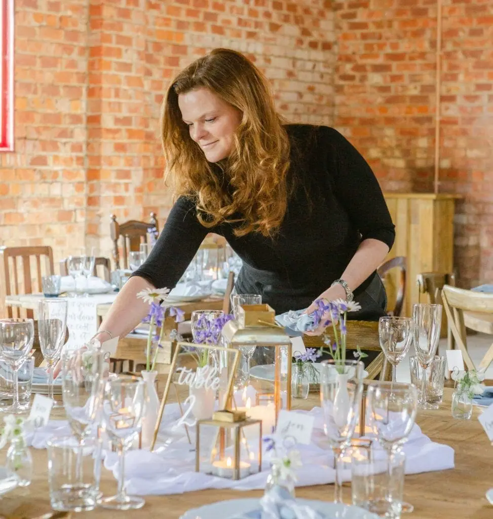 A woman arranges a table set with glasses, flowers, and candles in a brick-walled room.