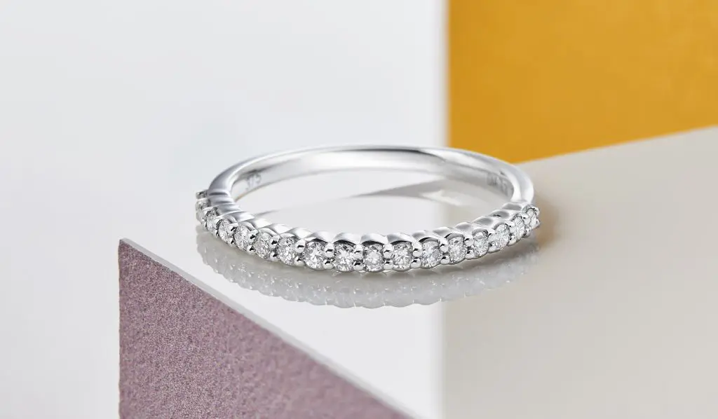 A silver ring with a row of small, sparkling diamonds is placed on a reflective surface with a minimalist background, perfect for showcasing in wedding services.