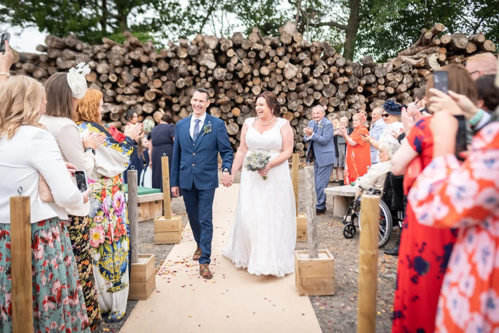 Bride and groom walk down outdoor aisle lined with guests and wooden logs, smiling and holding hands.