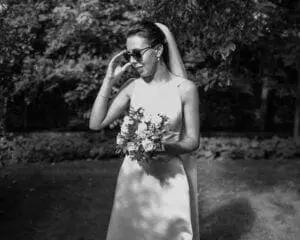 A bride in a white dress and veil stands outdoors, wearing sunglasses, and holding a bouquet of flowers, showcasing the elegance offered by premium wedding services.