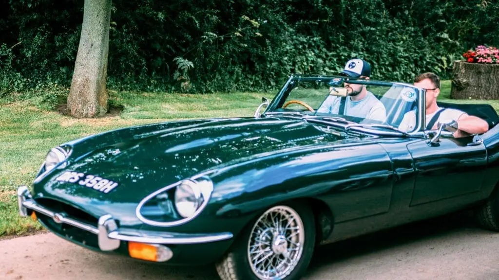 Two people are seated in a classic dark green convertible car with the top down, parked near a grassy area with trees and a flower pot, discussing wedding planning amidst the serene backdrop.