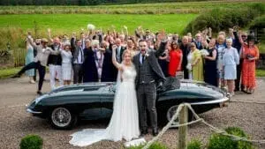 A bride and groom stand beside a vintage car with their hands raised, surrounded by a cheering group of well-dressed guests in an outdoor setting, celebrating their wedding UK style.