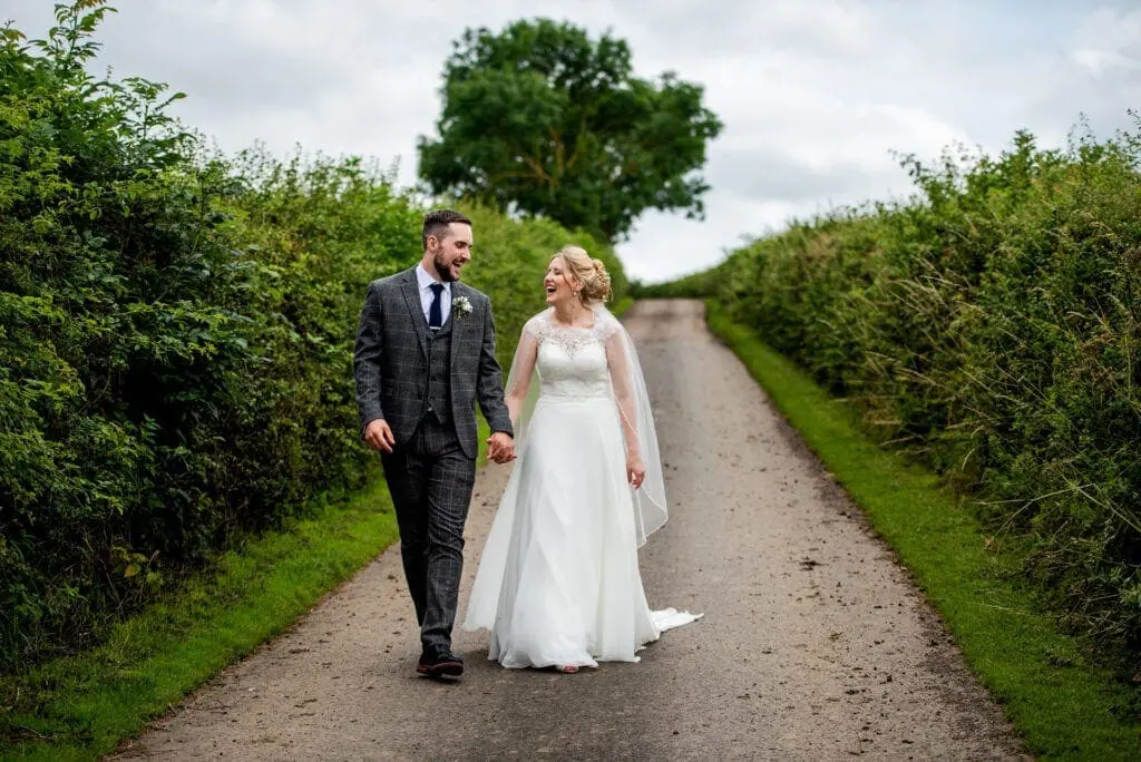 A couple in wedding attire walks hand in hand on a tree-lined path with grassy hedges, smiling at each other, embodying the joy and elegance of a perfectly planned wedding.