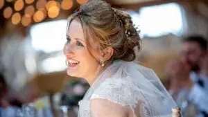 A bride in a white wedding dress and veil smiles while sitting at a table during a beautifully planned wedding reception. Blurred lights and guests are visible in the background, capturing the essence of meticulous wedding planning.