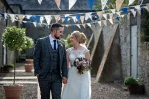 A bride and groom walk hand in hand, smiling at each other on their wedding day. The bride holds a bouquet, and they are outdoors beneath rows of decorative triangular flags—an enchanting scene that any wedding business would be proud to help plan.