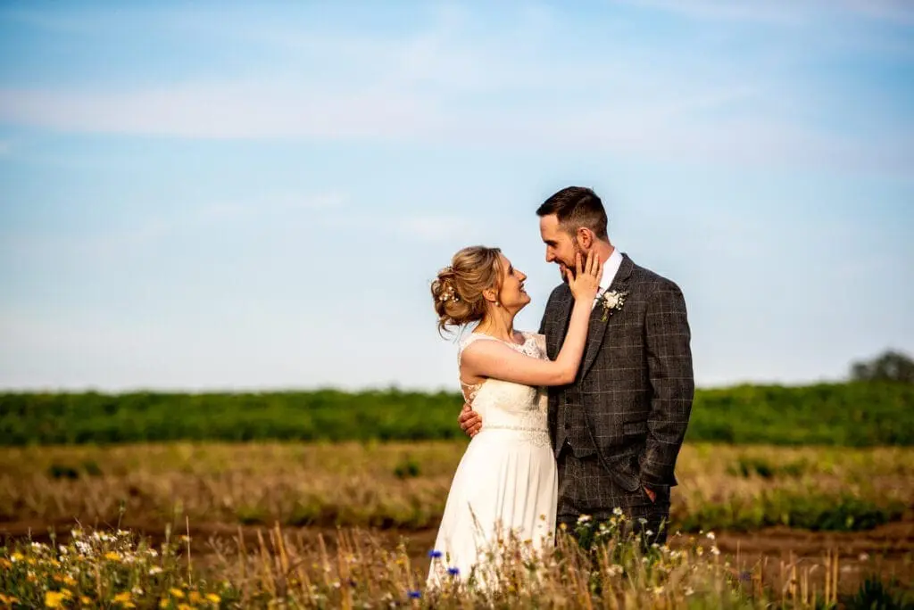 A bride and groom stand in a field with wildflowers, gazing at each other. The groom wears a gray plaid suit, and the bride wears a white dress, reflecting the charm of a classic wedding UK style.