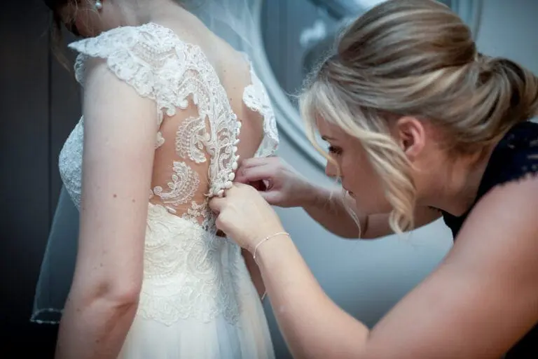 A woman in a dark dress is buttoning the intricate lace back of a white wedding gown worn by another woman, capturing a sweet moment in wedding planning.