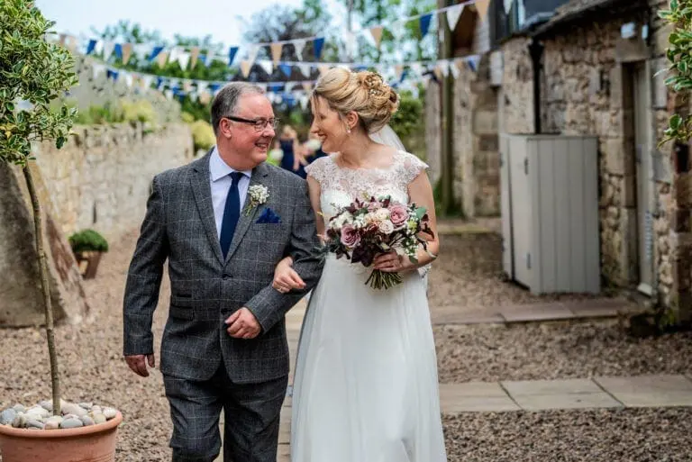 A bride in a white gown walks arm-in-arm with a man in a checkered suit down a rustic, outdoor path adorned with bunting. The bride holds a bouquet of flowers and they are both smiling, showcasing the flawless execution of expert wedding planning.