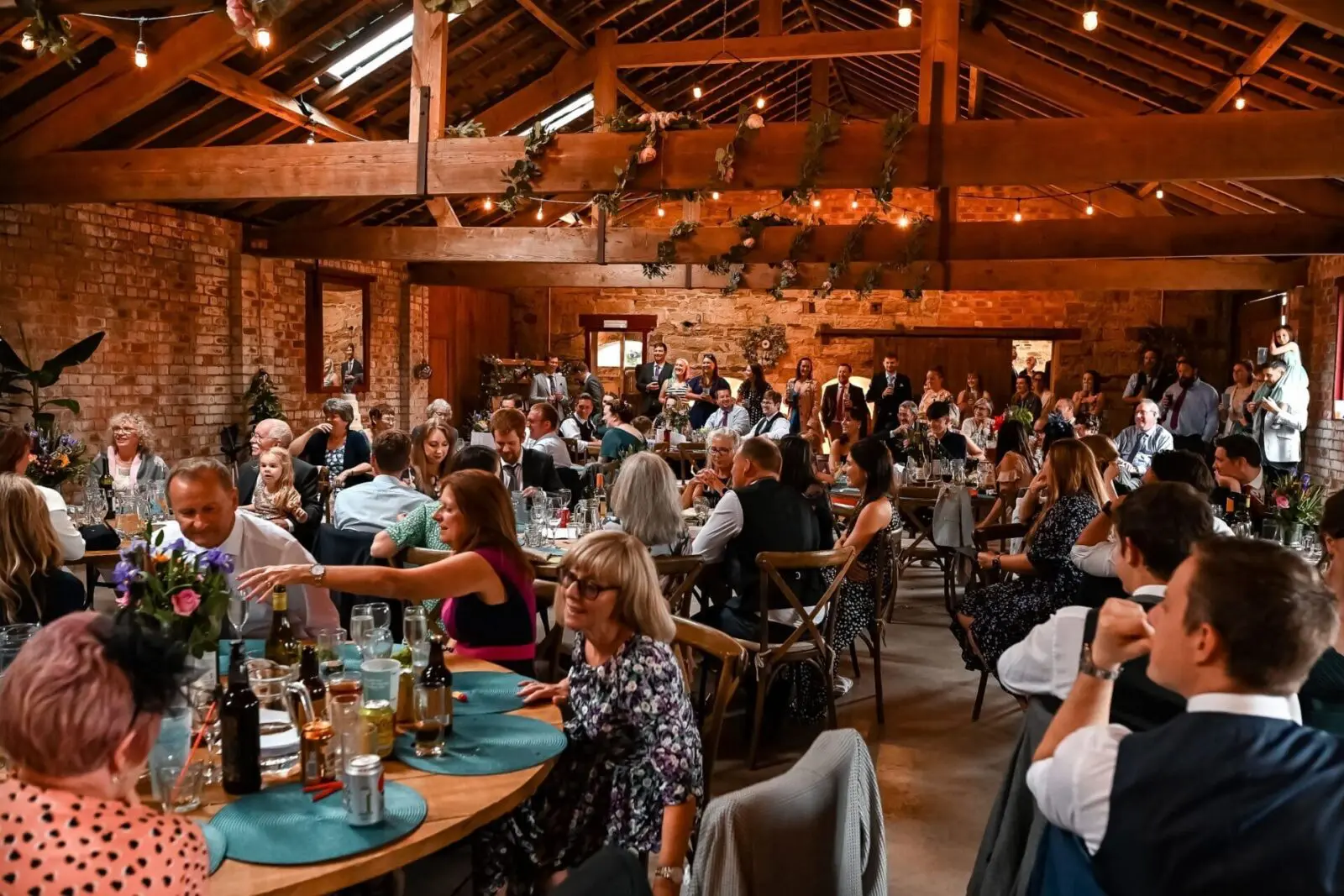 People gathered in a rustic venue with wooden beams, seated at tables decorated with flowers and drinks, attending an indoor event.