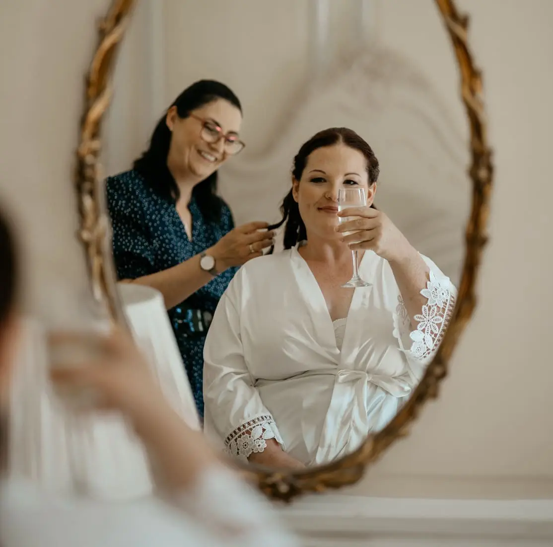 A woman in a robe smiles and holds a glass of white wine while another woman styles her hair, seen in an ornate mirror.