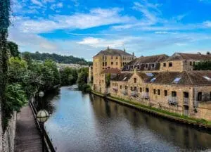 A serene river runs beside historic stone buildings under a partly cloudy blue sky, with lush greenery in the background—an idyllic setting perfect for a wedding.
