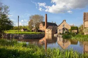 A serene rural scene featuring stone buildings by a calm pond, with ducks swimming, grassy areas, and tall trees under a partly cloudy sky; perfect for wedding planning in the UK.