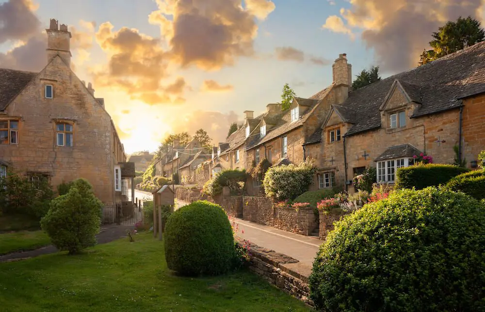 A picturesque village scene at sunset with stone houses, neatly trimmed hedges, and a tranquil atmosphere.