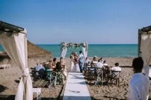 A wedding ceremony is taking place on a beach with an aisle leading to a floral arch. Guests are seated on either side, facing the bride and groom. The ocean and blue sky provide a stunning backdrop, a picturesque result of meticulous wedding planning.