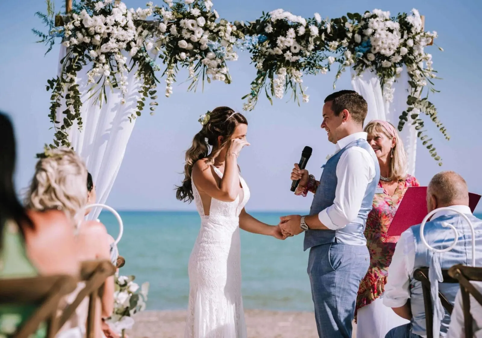 A bride and groom exchange vows under a floral arch at a beach wedding in the UK. The bride wipes her eyes while holding the groom's hand as he speaks into a microphone. Guests watch from their seated positions, savoring the beautifully planned ceremony.