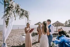 A couple stands under a floral arch on a beach, facing each other, while a woman officiates the wedding ceremony with a book in hand. Guests are seated and watching.